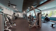 This single-story fitness center on 37th Street South in Saint Petersburg, Florida, offers a thoughtfully designed workout facility organized across multiple interconnected zones. The space is characterized by light aqua-toned walls that transition into deeper teal ombre accents, dark wood-paneled ceilings, recessed lighting, and ceiling fans throughout, creating a cohesive coastal ambiance.

The cardio zone features a row of treadmills, elliptical trainers, and StairMaster units positioned along large windows that frame views of tropical palm trees, flooding the area with natural light. Adjacent to this is a comprehensive strength training area equipped with cable machines, multi-station weight systems, benches, a functional training rig with TRX suspension systems, medicine balls, and a full dumbbell rack. Rubber flooring throughout provides durability and safety across all workout zones.

A dedicated cycling studio accommodates multiple stationary bikes and includes a wall-mounted television, decorative wall art, and additional ceiling fans, offering a bright and open environment for group or individual cycling sessions. A separate functional training room provides open floor space alongside wall-mounted storage for accessories including resistance bands, kettlebells, and stability balls, making it well-suited for dynamic, multi-modal workouts.
