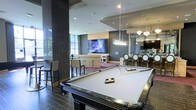 This stylish single-floor amenity space is designed for socializing and entertainment, combining modern aesthetics with a range of leisure features. At the forefront is a full-size billiards table finished in sleek black and white, positioned beneath recessed ceiling lighting that highlights the room's polished tile flooring. The space flows into a dedicated entertainment zone, where a large flat-screen television is mounted against a bold dark accent wall, flanked by high-top bistro tables and bar-height seating — ideal for gathering to watch events or simply unwind. Adjacent to the entertainment area is a well-appointed bar and lounge section, elevated with decorative pendant chandeliers that cast a warm, inviting glow over the bar stools and counter. Tall floor-to-ceiling windows along one side of the space flood the interior with natural light, creating an open, airy atmosphere while offering views of the surrounding greenery. The overall design palette draws on dark tones, warm neutrals, and contemporary finishes, resulting in a space that feels both sophisticated and welcoming. Every element is thoughtfully arranged to encourage both lively gatherings and relaxed downtime.