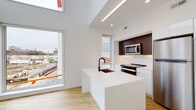 This contemporary two-floor space showcases a clean, minimalist design defined by crisp white walls, light hardwood flooring throughout, and an abundance of natural light streaming through large windows with urban views.

The open-plan kitchen serves as a central feature, anchored by a striking white waterfall island with an integrated sink and matte black faucet. Contrasting rich dark wood upper cabinetry pairs elegantly with white lower cabinets, while a full suite of stainless steel appliances — including a refrigerator, range, and built-in microwave — completes the space. Soaring double-height ceilings above the kitchen create a dramatic sense of volume, further enhanced by architectural linear lighting accents.

The living areas are bright and versatile, with expansive windows framing city views and generous floor plans ready for a variety of configurations. Recessed lighting supplements the natural light throughout.

The bathrooms are finished with floor-to-ceiling stone-look tile in warm neutral tones, featuring floating vanities with matte black fixtures, oversized mirrors, and walk-in shower areas, lending a refined, spa-like atmosphere.

A wood-tread staircase connects the two levels, and the upper floor offers additional rooms with their own windows and closet space, continuing the light and airy aesthetic found throughout the entire space.