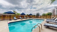 Located at 1240 Benchmark Park Dr in Columbus, Ohio, this outdoor amenity area is designed to offer residents a well-rounded recreational retreat. At the heart of the space is a generously sized outdoor swimming pool with crystal-clear blue water, framed by cobalt blue tile accents and accessible via stainless steel entry rails. The surrounding sundeck provides ample room for relaxation, lined with cushioned chaise lounge chairs and shaded by navy blue market umbrellas, making it an ideal spot for warm-weather enjoyment. A warm-toned wooden privacy fence encloses the pool area, creating a sense of seclusion while mature trees lend natural greenery and shade to the environment. Adjacent to the pool, a covered patio lounge area offers a comfortable gathering space furnished with wicker seating, accent chairs, and a central fire table — perfect for socializing in a relaxed outdoor setting. Lush potted plants and greenery enhance the ambiance of this lounge area. The multi-story residential building rises in the background, providing an attractive architectural backdrop. The overall design balances functionality and comfort, delivering a welcoming outdoor space suited for both active recreation and leisurely relaxation.