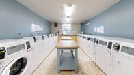Located at 150 Paisley in Mississauga, Ontario, this shared laundry facility is a practical and well-organized space designed to efficiently serve building residents. The room features rows of commercial-grade top-load and front-load washing machines and dryers lining both sides of the space, providing ample capacity for multiple users. Painted in a calming combination of soft blue and warm beige tones, the room maintains a clean and welcoming atmosphere. Centrally positioned folding tables with light wood surfaces and blue-grey frames offer convenient workspace for sorting and folding laundry. The terrazzo flooring is durable and easy to maintain, contributing to the room's overall tidiness. Overhead fluorescent lighting ensures the space is well-lit throughout. A window at the far end allows for natural light, enhancing the brightness of the room. A wall-mounted air conditioning unit provides climate control for year-round comfort. Additional amenities include a payment station, recycling and waste bins, posted instructional signage, and an electrical panel, all thoughtfully integrated to support a smooth and user-friendly laundry experience. The space is neatly maintained and thoughtfully laid out to maximize both functionality and ease of use.
