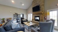 Located at 2753 Michaelsway Ave in Lancaster, Ohio, this single-floor space offers a warm and inviting atmosphere designed for comfort and gathering. The main living area commands immediate attention with its soaring vaulted ceilings that create a grand sense of openness and volume. The room's most striking architectural feature is a floor-to-ceiling stacked stone fireplace, which serves as a natural focal point and anchors the space with a rustic yet refined character. A wall-mounted flat-screen television above the fireplace mantel adds a modern, functional touch. The seating arrangement includes plush sofas and accent chairs in neutral tones, centered around a wood-top coffee table and layered area rug, encouraging relaxed conversation and lounging. An industrial-style open shelving unit provides both storage and decorative display. Natural light filters into the space through French doors that open to an outdoor area, further enhancing the airy feel. Recessed lighting in the ceiling complements the natural light, ensuring the space remains bright and welcoming at any hour. The overall aesthetic blends contemporary design with warm, natural materials, creating a balanced and comfortable environment.