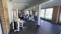 This single-floor fitness center offers a practical and well-organized workout environment designed to support a variety of strength training routines. The space is outfitted with multiple cable and weight machines, providing users with a comprehensive selection of resistance training equipment. Durable rubber flooring covers the workout area, offering both safety and comfort underfoot.

Full-length mirrors line one wall, enhancing the sense of space while allowing users to monitor their form during exercise. Structural columns are integrated throughout the floor plan, subtly defining the layout without disrupting the open feel of the room. Overhead, fluorescent lighting ensures the space is consistently and evenly illuminated.

Large windows along one side of the room invite an abundance of natural light and frame views of a well-maintained green outdoor area, creating a refreshing and motivating atmosphere. The combination of natural light, mirrored surfaces, and open floor space gives the gym an airy, spacious quality. The overall design is clean and functional, prioritizing ease of movement and accessibility to equipment, making it a well-suited facility for regular fitness use.