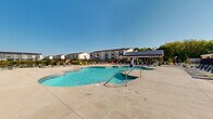 Located on Gusher Way in Lafayette, Indiana, this outdoor amenity space offers a well-appointed pool and recreation area designed for relaxation and leisure. At the heart of the space is a generously sized freeform swimming pool with a smooth, curved design and multiple entry points, its clear turquoise water complemented by a wide surrounding concrete deck. A covered pergola structure with a standing-seam metal roof anchors one side of the area, providing shaded gathering space alongside the pool. An adjacent building with large windows and a prominent metal roof serves as a clubhouse or amenity facility, offering interior views of the pool area from within.

The deck is outfitted with an extensive collection of lounge chairs, Adirondack-style seating in vibrant green, and modular outdoor furniture arrangements, creating numerous spots for sunbathing and socializing. The entire area is enclosed by a sleek black iron fence, with landscaped greenery and a scenic pond visible just beyond the perimeter, adding a natural backdrop to the setting. The surrounding residential buildings provide a sense of community scale. Under open skies, this pool area delivers a bright, welcoming outdoor environment ideal for warm-weather enjoyment.