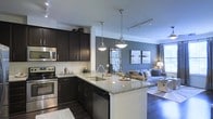 This single-floor space showcases a thoughtfully designed open-concept layout that seamlessly integrates the kitchen and living areas into one cohesive environment. The kitchen is a standout feature, outfitted with rich dark cabinetry, granite countertops, and a full suite of stainless steel appliances including a range, microwave, and dishwasher. A central island with an undermount sink doubles as a breakfast bar and prep space, illuminated by a pair of elegant pendant lights that hang from the ceiling. Track lighting above enhances the functional appeal of the kitchen. The transition into the living area is fluid and natural, with dark hardwood flooring running throughout and creating a warm contrast against the cool blue-grey accent wall. The living space is bright and welcoming, benefiting from natural light filtering through large windows dressed with curtain panels. Crown molding adds a refined architectural touch to the space. The overall ambiance is contemporary and polished, combining dark tones with lighter accents to create a balanced, stylish interior that feels both functional and inviting.