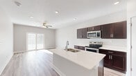 This well-designed single-floor unit offers a clean, modern aesthetic with a thoughtful layout that maximizes both function and comfort. The heart of the home is the open-concept kitchen and living area, where a large central island with a double-basin sink and quartz countertops serves as a natural gathering point. Rich dark-espresso cabinetry contrasts beautifully against the bright white counters, while a full suite of stainless steel appliances — including a side-by-side refrigerator with ice and water dispenser, electric range, built-in microwave, and dishwasher — provides a complete and contemporary kitchen experience. Light wood-look hard flooring extends throughout the main living areas, adding warmth and continuity. The living room is bright and airy, featuring recessed lighting, a ceiling fan, and sliding glass doors that open to an outdoor area, allowing generous natural light to fill the space. Bedroom areas are finished with plush carpeting, neutral-toned walls, and windows fitted with blinds. The bathroom includes a dark-cabinet vanity with quartz countertop, a tub-shower combination, and connects seamlessly to the rest of the unit via a hallway. Crisp white doors, clean baseboards, and a neutral color palette throughout give the space a fresh, move-in-ready feel.