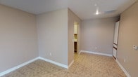 This single-floor space presents a straightforward, functional layout designed with simplicity and practicality in mind. The main area features neutral, warm greige walls that create a calm and versatile backdrop, complemented by crisp white baseboards that add a clean, finished look throughout. The flooring consists of tile laid in a classic pattern, providing durability and ease of maintenance.

The ceiling features a subtle angled element toward one corner, adding a modest architectural detail to the otherwise understated space. Recessed lighting is installed in the ceiling, providing focused illumination and contributing to an uncluttered overhead appearance. A visible HVAC vent ensures climate control is integrated directly into the ceiling design.

An open doorway leads to an adjoining room, visible in the background, which appears to contain cabinetry with darker wood tones — suggesting a utility, bathroom, or kitchenette function. The transition between spaces is seamless, maintaining the same neutral color palette throughout.

Overall, the space offers a clean, move-in-ready environment with a no-frills aesthetic, well-suited for a variety of uses. Its neutral tones and tile flooring make it highly adaptable for personalization.
