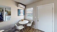 This single-floor unit offers a thoughtfully arranged layout that maximizes its compact footprint. The main living area features wood-look flooring and neutral gray walls beneath a textured ceiling, creating a clean, understated aesthetic. Multiple windows fitted with horizontal blinds allow ample natural light to fill the space. A wall-mounted mini-split AC unit ensures year-round climate control. A freestanding room divider screen provides a flexible way to define separate zones within the open layout, while a small round dining table with modern white chairs occupies a dedicated corner near the entry door.

The kitchenette is efficiently designed, featuring light granite-style countertops, a stainless steel double-basin sink, gray cabinetry above and below, and a pass-through window that connects visually to the living area. A dishwasher is integrated beneath the counter, adding practical convenience.

The bathroom is straightforward and functional, with a vanity featuring a speckle-finish countertop, a single-basin sink, gray cabinetry below, and a well-lit mirror above. The overall ambiance throughout the unit is modest and practical, with consistent neutral tones and durable finishes suited for everyday living.