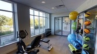 This fitness space is designed to support a well-rounded workout experience within a compact, efficiently organized layout. The room features large multi-pane windows along one wall, flooding the interior with natural light and offering views of the surrounding landscaping and trees. The neutral gray walls and drop-tile ceiling contribute to a clean, functional aesthetic, while the low-pile carpet flooring adds a degree of comfort underfoot.

The space is equipped with a stationary exercise bike and an adjustable workout bench, catering to both cardiovascular and strength training needs. A tiered dumbbell rack holds a range of free weights, making the area suitable for varied resistance training routines. A collection of stability and exercise balls in multiple sizes is stored on a dedicated rack, supporting core and flexibility workouts.

Glass-paneled sliding doors lead to an adjacent room visible in the background, which appears to extend the facility's functional footprint. Informational signage is mounted near the entrance, and the overall layout promotes ease of movement between equipment. The space strikes a balance between practicality and comfort, making it an accessible fitness option.