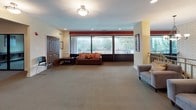 This single-floor community space is thoughtfully designed to serve as a versatile gathering and recreation hub. The main lounge area makes an immediate impression with its expansive layout, neutral carpeting, and large floor-to-ceiling windows that frame views of the surrounding landscape and an adjacent outdoor balcony. A bold red accent band running along the upper wall adds a vibrant contrast to the warm yellow and cream tones throughout. Comfortable seating, including a brown sofa with orange accent pillows and a pair of plush gray armchairs, is arranged to encourage relaxed conversation, while elegant pendant chandeliers and recessed lighting illuminate the space warmly. A textured white brick feature wall adds architectural character near the seating nook, complemented by a metal railing that defines the lounge's open-plan layout. The activity area transitions seamlessly from the lounge and offers a flexible layout with folding tables and colorful chairs, a large flat-panel display, and a pool table positioned in the background — ideal for both casual recreation and organized gatherings. Framed artwork in earthy tones is displayed throughout, contributing to the inviting, community-oriented ambiance. Dark wood doors provide clean transitions between areas, completing a polished and functional space suited for a range of social activities.