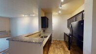 This single-floor residence offers a well-organized layout that balances functional living spaces with modern finishes. The kitchen serves as a central highlight, featuring dark espresso cabinetry, granite countertops with a light speckled pattern, and a peninsula with a stainless steel sink that opens toward the main living area. Black appliances — including a range, microwave, and refrigerator — complement the cabinetry, while warm wood-look flooring ties the space together. Track lighting illuminates both the kitchen and adjacent areas effectively.

The open living area transitions between wood-look hard flooring and plush carpet, creating a natural distinction between zones. Track lighting continues here, providing ample illumination, and the space offers a clean, neutral palette throughout.

The bedroom is carpeted and features a globe-style chandelier as a decorative focal point, along with a window that allows natural light to enter. An adjacent bathroom includes a granite-topped vanity with dark cabinetry, a large mirror with vanity lighting, and direct access to a spacious walk-in closet fitted with wire shelving systems.

A dedicated laundry area houses a full-size Whirlpool top-loading washer and dryer, accompanied by additional cabinetry for storage. The overall ambiance is clean, contemporary, and move-in ready.