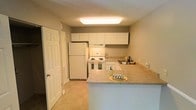 This two-story residence offers a practical and well-organized living space. The kitchen is a standout area, designed with functionality in mind. White raised-panel cabinetry lines the walls, providing ample storage, while the layout incorporates a peninsula with a light-toned countertop that doubles as a breakfast bar, creating a natural division between the kitchen and adjacent living areas. White appliances, including a full-size refrigerator and an electric coil range with an overhead exhaust, complement the cabinetry for a clean, cohesive look. A sink is positioned along the far wall, completing the efficient work triangle. Light tile flooring runs throughout the kitchen, contributing to the bright and easy-to-maintain aesthetic. Overhead, a large flush-mount fluorescent light fixture provides even illumination across the space. The neutral taupe wall color lends a warm yet understated tone throughout. A paneled interior door near the kitchen suggests access to additional rooms or storage. The two-floor layout indicates additional living, sleeping, or utility spaces on the second level, expanding the overall footprint of this residence beyond what the kitchen image alone conveys.