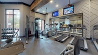 Located in Thompson's Station, Tennessee, this single-story fitness center at 2214 Vintage Tollgate Drive offers a thoughtfully designed workout facility that balances functionality with refined aesthetics. The space features soaring ceilings accented by rustic wood beams, which contrast attractively against the neutral-toned walls, creating a modern yet inviting atmosphere.

The fitness floor is well-appointed with a comprehensive selection of equipment, including multiple treadmills, elliptical machines, stationary cycles, and a cable strength-training system, alongside free weights and benches. Large floor-to-ceiling mirrors line portions of the walls, amplifying the sense of space and allowing users to monitor their form during workouts. Wall-mounted flat-screen televisions are positioned for visibility across the floor, adding convenience during cardio sessions.

The flooring combines tile and modular carpet sections, providing both durability and comfort underfoot. Recessed lighting and pendant fixtures ensure the space is well-lit throughout. A glass-paneled entry connects the fitness area to an adjacent room featuring decorative artwork and a chandelier, adding an upscale touch. Natural light enters through exterior-facing doors, contributing to the bright, energetic ambiance of the overall space.