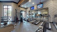 Located in Thompson's Station, Tennessee, this single-story fitness center at 2214 Vintage Tollgate Drive offers a thoughtfully designed workout facility that balances functionality with refined aesthetics. The space features soaring ceilings accented by rustic wood beams, which contrast attractively against the neutral-toned walls, creating a modern yet inviting atmosphere.

The fitness floor is well-appointed with a comprehensive selection of equipment, including multiple treadmills, elliptical machines, stationary cycles, and a cable strength-training system, alongside free weights and benches. Large floor-to-ceiling mirrors line portions of the walls, amplifying the sense of space and allowing users to monitor their form during workouts. Wall-mounted flat-screen televisions are positioned for visibility across the floor, adding convenience during cardio sessions.

The flooring combines tile and modular carpet sections, providing both durability and comfort underfoot. Recessed lighting and pendant fixtures ensure the space is well-lit throughout. A glass-paneled entry connects the fitness area to an adjacent room featuring decorative artwork and a chandelier, adding an upscale touch. Natural light enters through exterior-facing doors, contributing to the bright, energetic ambiance of the overall space.