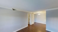 This single-floor space presents a clean, understated aesthetic defined by warm hardwood flooring and soft, neutral gray walls that create a calm, versatile backdrop. Crown molding runs along the ceiling perimeter, adding a touch of classic architectural detail to the otherwise straightforward design. The main living area is generously proportioned, offering an open feel well-suited for a variety of furniture arrangements.

A mirrored sliding closet provides both practical storage and the visual effect of expanded space, reflecting light throughout the room. An elegant chandelier anchors the far end of the room, contributing warmth and a refined finishing touch to the ceiling.

A doorway visible from the main area leads to an adjacent space, likely a bathroom or utility room, identifiable by its louvered door and interior shelving. Central air vents integrated into the ceiling and walls indicate a full HVAC system throughout.

White baseboards and trim provide crisp contrast against the darker flooring, reinforcing the space's tidy, well-maintained character. Overall, the space offers a bright, neutral canvas ready for personalization.