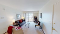 This single-floor residence offers a thoughtfully arranged layout across several well-defined spaces. The living room serves as the heart of the home, featuring plush wall-to-wall carpeting, neutral walls, and a large sliding glass door flanked by vertical blinds that frames outdoor views of natural wooded scenery while flooding the room with natural light. Framed artwork accents the walls, adding character to the bright, open atmosphere.

The galley-style kitchen is efficiently designed with dark wood cabinetry lining both walls, granite-toned countertops, wood-look flooring, and white appliances including a gas range and refrigerator, providing ample storage and workspace.

The primary bedroom is a comfortable retreat with carpeted flooring, a ceiling fan with light fixture, and a window with blinds offering soft natural light. A second bedroom mirrors this layout with its own window and cozy proportions.

The bathroom features a shower/tub combination with a decorative curtain and neutral finishes, completing the unit's functional design. Throughout, the residence maintains a clean, understated aesthetic with consistent neutral tones and practical finishes.