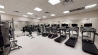 This fitness center offers a spacious, thoughtfully designed workout environment housed on a single floor. The room features a drop ceiling with large recessed panel lighting that bathes the space in bright, even illumination, creating an energetic and focused atmosphere conducive to exercise.

The cardio section is prominently featured, with a row of treadmills positioned at the forefront, each equipped with individual monitor screens for entertainment during workouts. Behind them, a selection of elliptical trainers and additional cardio machines provides variety for users with different training preferences. The strength and cross-training equipment is arranged further into the space, allowing for a logical and efficient flow throughout the facility.

Full-length mirrors line the walls, visually expanding the room while allowing users to monitor their form during exercise. The light-toned flooring is clean and durable, suited for high-traffic fitness use. HVAC vents are integrated into the ceiling, ensuring consistent climate control throughout.

The overall layout is open and organized, providing ample space between equipment for safe and comfortable movement. The clean, neutral color palette and modern equipment give the facility a professional and welcoming character, making it well-suited for a range of fitness levels and training routines.