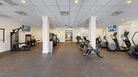 Located at 1111 W Bay Ave in Barnegat, NJ, this fitness center offers a generously sized, single-floor facility designed to support a full range of workout routines. The space is thoughtfully organized into distinct training zones, providing a comprehensive exercise environment for users of all fitness levels.

The cardio area features multiple treadmills, elliptical machines, stationary bikes, a rowing machine, and a stair climber, all arranged for comfortable spacing between equipment. A wall-mounted flat-screen TV accompanies this zone, enhancing the workout experience. Spin bikes occupy an open central area, ideal for cycling sessions.

The strength training section is well-appointed with a dumbbell rack, cable machines, a weight bench, and medicine balls and kettlebells, offering versatility for both free-weight and machine-based workouts.

Architecturally, the space features a drop-panel ceiling with recessed lighting and ceiling fans that ensure even illumination and airflow throughout. Decorative white columns add a classic character to the interior. Rubber flooring provides safety and durability across the entire space. Large windows with striped valances and glass-paneled double doors allow ample natural light and views to the outdoors. The clean white walls and bright, open layout create a welcoming and motivating atmosphere.