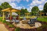 Located at 100 Highland Park Trail in Sandy Springs, Georgia, this thoughtfully designed outdoor amenity space offers a welcoming retreat set among mature trees and lush greenery. The grounds are anchored by multiple wooden pergolas that provide shaded gathering areas, furnished with wicker egg chairs featuring bold red cushions and sectional seating — perfect for relaxing or socializing outdoors.

At the heart of the space, a stone fire pit surrounded by natural flagstone hardscaping serves as a central focal point, ideal for evening gatherings. Neatly manicured garden beds run through the space, planted with a diverse mix of ornamental grasses, shrubs, and flowering plants set against a backdrop of rich pine mulch. Wide concrete pathways connect the various seating areas and pergola structures, ensuring easy navigation throughout the grounds.

Well-maintained open lawn areas provide flexible green space, while additional pergola structures are positioned throughout to create distinct outdoor rooms within the larger landscape. The perimeter is bordered by established trees and layered plantings that offer privacy and a natural, park-like ambiance. The overall design balances structured landscaping with comfortable, inviting furnishings, creating a serene outdoor environment suited for both quiet relaxation and community gatherings.