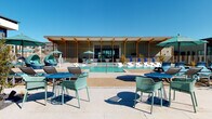 This expansive outdoor amenity space is designed around a centrally positioned swimming pool that serves as the focal point of the entire deck. The pool area is flanked by a generous sundeck lined with cushioned lounge chairs, offering ample space for relaxation under open skies. The surrounding hardscape combines smooth concrete pavers and aggregate-finished surfaces, creating distinct zones for lounging, dining, and socializing.

A contemporary single-story clubhouse structure anchors one end of the pool deck, featuring a distinctive pergola-style facade with exposed timber columns and large-format glazing that blends indoor and outdoor spaces seamlessly. Additional covered pergola structures along the perimeter provide shaded retreats equipped with teal and blue outdoor dining sets and bistro tables arranged for casual gatherings.

A dedicated lounge nook, sheltered beneath a sail-shade canopy, offers plush sectional seating with colorful accent pillows and a round coffee table—ideal for relaxed conversation. Potted plants and landscaped gravel beds introduce natural texture throughout the space. The overall aesthetic is modern and resort-inspired, combining clean architectural lines with vibrant teal and blue furnishings set against an open, sun-drenched sky.