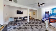 Located at 202 Onley Rd in Salisbury, Maryland, this community game room is a single-floor recreational space designed for social activity and entertainment. The open-plan layout is anchored by patterned carpet tiles in a modern geometric design, lending the space a contemporary, energetic aesthetic throughout.

The room is equipped with an impressive array of gaming options. Arcade machines, including skee-ball and a basketball shooter, occupy one section of the space, while a foosball table and shuffleboard table provide additional entertainment. Multiple wall-mounted flat-screen televisions are positioned throughout for viewing convenience. Ceiling fans are installed across the space, ensuring comfortable airflow.

Seating is thoughtfully distributed around the room in the form of high-top bistro tables paired with bar-height chairs, offering casual spots for guests to gather and socialize. Structural white columns define zones within the open floor plan without obstructing sightlines or flow between areas.

Natural light enters through several white-framed windows, offering views of lush surrounding greenery. The white walls and textured ceilings keep the atmosphere bright and clean, complementing the vibrant colors of the gaming equipment. A restroom is accessible from within the space. Overall, the room delivers a lively, welcoming environment well-suited for community recreation.