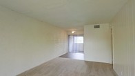 This single-story space features a straightforward, functional layout highlighted by a open living area. The room is finished with wall-to-wall neutral-toned carpet, providing a soft underfoot feel throughout. The textured ceiling adds a classic architectural detail, while a centrally mounted ceiling light fixture provides overhead illumination. A large window dressed with vertical blinds serves as the room's primary source of natural light, allowing daylight to filter into the interior. The walls feature a subtle paneled texture on one side, adding a modest design element to the otherwise clean, neutral palette. An overhead HVAC vent is visibly integrated into the upper wall, ensuring climate control throughout the space. The overall ambiance is calm and understated, offering a blank canvas well-suited for personalization and furnishing.