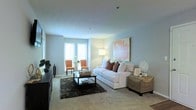 This single-floor space showcases a thoughtfully designed living area that balances comfort and style. The room is finished with soft wall-to-wall carpeting and painted in a light, neutral grey-blue tone that contributes to a calm and airy atmosphere. Natural light filters in through two windows positioned in the corner of the room, illuminating a secondary seating area furnished with a pair of warm orange accent chairs and a delicate glass-top side table — a charming spot ideal for reading or conversation.

The main seating area centers around a plush white sofa accented with richly toned decorative pillows, anchored by a dark rectangular coffee table atop a textured dark area rug. A woven storage ottoman adds both function and texture nearby. A large framed abstract artwork in blush and gold tones serves as a focal point on the wall, complementing the warm accents throughout the room.

A media console along one wall provides additional storage and display space. Overhead, the smooth ceiling features subtle recessed lighting. The overall ambiance is inviting and well-curated, blending neutral foundations with warm accent tones throughout.