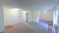 This single-floor residential space offers a straightforward, functional layout designed for comfortable everyday living. The main living area is generously sized, finished with wall-to-wall carpeting that adds warmth underfoot. Smooth, light-toned walls create a clean, neutral backdrop throughout, while ceiling-mounted light fixtures provide consistent illumination across the space.

A partial wall divides the living area from the rest of the floor plan, adding a subtle sense of structure without fully closing off the open feel. A built-in closet with bi-fold doors is conveniently positioned along this dividing section, offering practical storage directly accessible from the living space.

Adjacent to the main living area, the kitchen is visible and features overhead cabinetry, a countertop workspace, and white appliances, providing a functional cooking environment. The transition between the carpeted living area and the kitchen space is clearly defined, maintaining a logical flow between the two zones.

The overall ambiance is bright and understated, with a neutral color palette that offers a versatile canvas for personalization. The single-floor layout ensures easy navigation throughout, making this a practical and accessible living space.