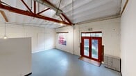 Located at 2888 E Walnut St in Pasadena, CA, this two-floor loft-style space blends industrial character with functional versatility. The expansive main floor is anchored by a dramatic vaulted ceiling with exposed red-painted timber trusses and visible ductwork, lending a raw, warehouse aesthetic. Painted white brick walls, smooth light-grey concrete floors, and track lighting create a bright, open-plan environment ideal for both living and working. Bold red-framed double glass entry doors welcome natural light into the generous main living area, which flows seamlessly through connected rooms. A staircase with a metal handrail leads to the upper mezzanine level, which features warm hardwood floors, sloped ceilings that follow the roofline, and exposed structural beams — offering a more intimate, cozy atmosphere in contrast to the main floor's volume. Two bathrooms serve the space: one is a compact, utilitarian powder room with dark tile flooring, a wall-mounted sink, and a round mirror; the other is a well-appointed full bathroom featuring a soaking tub, white tile with decorative blue and green accents, a floating vanity, and a large mirror. A hallway connects key areas of the upper level. The overall ambiance is that of a creative, industrial-chic space with character-rich architectural details throughout.