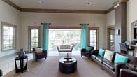 This single-story clubhouse offers a thoughtfully designed communal lounge that balances comfort and style. The main gathering area is anchored by a seating arrangement that includes dark leather sofas and armchairs paired with a tufted leather ottoman as a central coffee table, creating an inviting environment for socializing or relaxing. Crown molding along the upper walls adds an architectural refinement to the otherwise clean, neutral interior.

Natural light is a defining feature of this space, with a large central opening flanked by two glass-paned doors that lead directly to an outdoor patio or terrace. Floor-length teal curtains frame the openings, introducing a pop of color that complements the neutral beige walls and carpeted flooring. The outdoor area beyond is visible through the expansive opening, offering views of greenery and landscaping that enhance the sense of openness within the interior.

Recessed ceiling vents ensure climate comfort throughout the space, while emergency exit signage reflects the building's practical considerations. The overall ambiance is warm, airy, and well-appointed, making it a functional and attractive gathering space for residents.