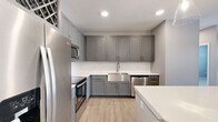 This single-floor unit showcases a thoughtfully designed, contemporary interior with a cohesive neutral color palette throughout. The kitchen serves as a standout feature, equipped with gray shaker-style cabinetry extending floor to ceiling, white quartz countertops, a stainless steel farmhouse sink, and a full suite of stainless steel appliances including a refrigerator, range, microwave, and dishwasher. A built-in wine rack adds a distinctive decorative touch. A central island with pendant lighting provides additional prep space and creates an open, functional layout. Light wood-look flooring runs throughout the main living and kitchen areas, contributing to a clean, modern aesthetic.

The living area is bright and open, featuring a glass-panel entry door with a transom window and an adjacent full-size window, allowing natural light to fill the space. Bedrooms are carpeted and feature large windows with outdoor views, providing a comfortable, quiet retreat.

The bathroom is well-appointed with a gray vanity, white quartz countertop, a large framed mirror, and a glass-enclosed walk-in shower with marble-style tile surround. A stacked washer and dryer unit is conveniently located within the bathroom, alongside access to a walk-in closet with built-in shelving.