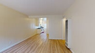 Located at 15003 Lemoli Ave in Gardena, California, this single-floor unit offers a clean, functional layout with a bright and open feel throughout. Light wood laminate flooring runs consistently across the main living areas, contributing to a cohesive and airy aesthetic. The living area is generously sized with neutral cream-toned walls, a wall-mounted heating and ventilation unit, and an entry door that opens directly into the space. Natural light filters in through large windows fitted with vertical blinds, keeping the interior well-lit. A wide, open corridor connects the living area to the kitchen, which features white cabinetry, dark countertops, a mosaic tile backsplash, and a gas range. The kitchen also benefits from a large window with vertical blinds, allowing ample natural light to fill the cooking space. The bathroom is tiled in clean white square tiles from floor to ceiling, with a combination soaking tub and shower, a sliding frosted privacy window above the tub, and a white vanity with cabinet storage. The overall design of the unit is practical and well-maintained, with a neutral color palette that provides a versatile canvas throughout.
