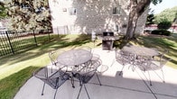 This outdoor patio space offers a welcoming and functional area designed for communal gathering and outdoor dining. The patio is finished with a smooth concrete surface and is furnished with classic wrought iron tables and chairs, including multiple seating arrangements that can accommodate groups of varying sizes. A full-sized gas grill station is positioned conveniently on the patio, making the space well-suited for outdoor cooking and entertaining. The surrounding landscape features a well-maintained grassy lawn shaded by mature trees, lending a relaxed, park-like atmosphere to the area. A decorative iron fence borders part of the perimeter, providing a sense of enclosure while maintaining an open feel. The adjacent brick building provides a sturdy architectural backdrop, with ground-level windows adding a sense of connection between the interior and exterior. Natural light filters through the tree canopy, creating a pleasant, dappled environment throughout the day. Overall, this patio area combines practical amenities with comfortable outdoor furnishings, making it an inviting space for casual gatherings, al fresco dining, and leisurely outdoor enjoyment.