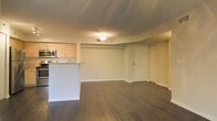 This single-floor space features an open-concept layout that integrates the kitchen and living area into one cohesive environment. The dark hardwood flooring runs throughout, providing a rich contrast against the light-toned walls and creating a grounded, contemporary aesthetic.

The kitchen is positioned to one side and is equipped with light maple cabinetry, stainless steel appliances including a microwave and range, and a peninsula counter with a light stone-style surface. The peninsula serves as a natural divider between the kitchen and the open living area, offering both functional workspace and an informal dining or gathering spot.

The living area is generously proportioned, offering flexibility for various furniture arrangements. A textured ceiling adds subtle visual interest overhead, while flush-mounted ceiling lighting provides consistent illumination throughout the space. The entry door is visible along one wall, framed cleanly within the layout.

Overall, the space presents a clean, functional design with a neutral palette, making it highly adaptable. The open flow between the kitchen and living areas enhances the sense of space, while the dark flooring anchors the interior with a modern, polished character.