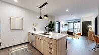 This single-floor residence is designed with a clean, contemporary aesthetic that emphasizes open living and natural light. The open-concept kitchen serves as a central hub, anchored by a large island with a white quartz countertop, an undermount sink with a matte black faucet, and light wood cabinetry with black hardware. Dual pendant lights hang above the island, while stainless steel appliances and a subway tile backsplash complete the functional, stylish space. The kitchen flows seamlessly into the living area, which benefits from floor-to-ceiling sliding glass doors leading to a private balcony. The outdoor space features adecked floor and open views of the surrounding urban landscape. The bedrooms are bright and tranquil, each featuring large picture windows that frame city views and allow generous natural light. Soft carpeting, clean white walls, and warm wood furnishings reinforce the unit's cohesive modern palette. The bathroom is finished with a light wood vanity, white quartz countertop, matte black fixtures, a large framed mirror, and a tiled tub-shower combination. Wood-look plank flooring runs throughout the main living areas, tying the space together with warmth and consistency.