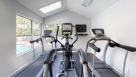 This well-equipped fitness center at 128 Chestnut Ridge Dr in Harrisonburg, Virginia, offers a comprehensive workout environment spread across a single floor. The space is thoughtfully organized into distinct exercise zones, each designed to support a variety of fitness routines. The cardio area features elliptical trainers, treadmills, a stationary bike, and a rowing machine, positioned beneath vaulted ceilings with large windows that invite natural light and provide pleasant outdoor views of surrounding greenery. A wall-mounted television adds entertainment for cardio sessions. The strength training section is equipped with a multi-station cable machine, a full rack of kettlebells, dumbbells, exercise balls, foam rollers, and workout mats, all neatly organized on storage racks. Floor-to-ceiling mirrors in the weight area enhance the sense of space and allow for proper form checks during exercise. Throughout the facility, durable rubber flooring provides safety and comfort underfoot. Recessed and flush-panel fluorescent lighting ensures consistent, bright illumination across all areas. The light blue and white walls contribute to a clean, motivating atmosphere. A water cooler, wall clock, and accessible doorways round out the practical amenities, making this fitness center a fully functional and welcoming space for residents.