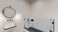 Located at 200 W Main St in New Albany, Ohio, this single-floor space showcases a thoughtful blend of classic and functional design. The most visually striking feature visible is the bathroom, which exemplifies timeless craftsmanship. The shower area is lined with crisp white subway tiles accented by dark grout, creating a clean yet striking contrast. The shower floor features charcoal hexagonal mosaic tiles, adding texture and visual depth. A clear glass panel enclosure maintains an open, airy feel within the space. Vintage-inspired bronze hardware and a traditional wall-mount shower fixture contribute to the room's character and charm. A wall-mounted sconce with a classic globe shade provides warm, ambient lighting, while a round brass-framed mirror completes the refined aesthetic. The porcelain wall-mount sink with navy blue trim reinforces the vintage sensibility throughout. Crisp white walls and clean trim work lend the space a bright, well-maintained appearance. Overall, the design reflects a careful attention to detail, pairing period-appropriate fixtures with durable, high-quality materials to create a space that is both functional and visually cohesive.
