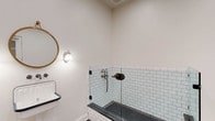 Located at 200 W Main St in New Albany, Ohio, this single-floor space showcases a thoughtful blend of classic and functional design. The most visually striking feature visible is the bathroom, which exemplifies timeless craftsmanship. The shower area is lined with crisp white subway tiles accented by dark grout, creating a clean yet striking contrast. The shower floor features charcoal hexagonal mosaic tiles, adding texture and visual depth. A clear glass panel enclosure maintains an open, airy feel within the space. Vintage-inspired bronze hardware and a traditional wall-mount shower fixture contribute to the room's character and charm. A wall-mounted sconce with a classic globe shade provides warm, ambient lighting, while a round brass-framed mirror completes the refined aesthetic. The porcelain wall-mount sink with navy blue trim reinforces the vintage sensibility throughout. Crisp white walls and clean trim work lend the space a bright, well-maintained appearance. Overall, the design reflects a careful attention to detail, pairing period-appropriate fixtures with durable, high-quality materials to create a space that is both functional and visually cohesive.