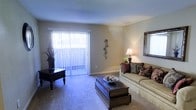 This single-floor residence offers a welcoming and well-appointed living environment designed for everyday comfort. The living room serves as the heart of the space, featuring plush wall-to-wall carpeting that adds warmth underfoot. A large sliding glass door dressed with vertical blinds floods the room with natural light while providing direct access to the outdoors. Neutral-toned walls create a versatile backdrop that complements a range of furnishing styles. The room is thoughtfully arranged with a cushioned sofa accented by decorative pillows, a dark wood trunk-style coffee table, and a matching side table that adds a classic touch. A framed wall mirror and a table lamp with a warm glow enhance the inviting ambiance, while decorative wall art and indoor greenery add personality and life to the space. A secondary window on the adjacent wall further contributes to the bright, airy feel of the room. The overall atmosphere is calm and residential, combining functional layout with comfortable, homey aesthetics that make this a practical yet charming living space.