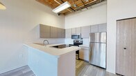 Located at 550 W Western Ave in Muskegon, Michigan, Unit 327 is a thoughtfully designed loft-style space that masterfully balances industrial heritage with modern updates. The open-concept layout is anchored by soaring ceilings with exposed wooden beams and visible ductwork, lending an authentic warehouse aesthetic throughout.

The kitchen is a standout feature, outfitted with sleek, flat-front cabinetry in a warm taupe finish, white quartz countertops, and a full suite of stainless steel appliances including a refrigerator, electric range, built-in microwave, and dishwasher. A peninsula with an integrated sink provides additional prep space and a natural divider between the kitchen and living area.

The living and bedroom areas showcase plush carpet flooring and are distinguished by exposed brick accent walls and wood-trimmed windows that allow natural light to filter in, reinforcing the loft's character. Wood plank-style flooring runs through the kitchen and entry areas, complementing the overall warm, industrial palette.

Two full bathrooms are finished with light wood-look tile flooring, modern floating vanities with white countertops, and full tub-shower combinations, offering clean, functional spaces. Exposed wooden posts appear throughout, tying the historic architectural elements cohesively into the unit's contemporary design.