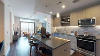 This well-appointed one-bedroom, one-bathroom residence showcases a thoughtfully designed open-concept floor plan with modern finishes throughout. The kitchen serves as a central feature, outfitted with light wood cabinetry, under-cabinet LED lighting, a textured tile backsplash, stainless steel appliances including a gas range and French door refrigerator, and a spacious island with a dark quartz countertop and bar seating. Pendant lighting adds a refined touch overhead, while rich dark hardwood flooring anchors the space.

The living area offers generous room for seating and opens directly onto a private balcony through large sliding glass doors, framing sweeping city skyline views. Natural light flows freely throughout the main living spaces.

The bedroom is a calm retreat with plush carpeting, a ceiling fan, and a large window showcasing urban panoramas. It connects directly to the bathroom and a walk-in closet with built-in shelving. The bathroom features a dual-sink vanity with dark cabinetry, a full soaking tub, and floor-to-ceiling gray tile accents, all finished with clean, contemporary tile flooring.
