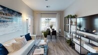 This single-floor residence offers a thoughtfully designed open-concept layout with consistent wood-look flooring and a neutral color palette throughout. The living and dining area flows seamlessly together, anchored by large windows dressed with patterned curtains that bring in ample natural light. A wall-mounted TV, open shelving units, and a glass-top coffee table complement the relaxed yet functional aesthetic.

The kitchen is well-equipped with warm-toned wood cabinetry, mosaic tile backsplash, stainless steel appliances including a refrigerator, range, and over-the-range microwave, plus generous counter space. A peninsula with seating for two offers a casual dining option in addition to the adjacent dining area.

The primary bedroom features sliding glass doors opening to a private balcony with outdoor seating, providing pleasant exterior views. A second bedroom is similarly outfitted with balcony access, while a third bedroom offers carpeted flooring and a large window. Both bathrooms include tub-shower combinations, wood-look flooring, and clean, neutral finishes. The overall ambiance is modern, light, and practical.