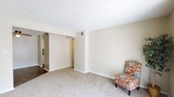 This single-floor space presents a clean, functional layout centered around a carpeted living area finished in warm, neutral tones. The walls are painted in a soft beige, creating a calm and understated ambiance throughout. Underfoot, light-colored wall-to-wall carpeting adds a sense of warmth and comfort to the main living space. The textured popcorn ceiling runs uniformly across the room, and a ceiling vent indicates a central HVAC system for climate control. A wide, open passageway connects the living area to an adjoining hallway and additional rooms beyond, suggesting a flowing, interconnected floor plan. The transition from carpet to hardwood or laminate flooring is visible through the passage, hinting at a distinct change in room function further in. White baseboards frame the perimeter of the room, offering a crisp contrast to the neutral walls. An electrical outlet is visible along the base of the main wall, and the overall layout appears practical and versatile. The space is open and airy, offering a blank canvas well-suited for a variety of furniture arrangements and interior styling preferences.