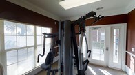 This fitness room offers a dedicated space for strength and conditioning, anchored by a large multi-station cable and weight machine that supports a wide variety of exercises. The room is finished with classic crown molding along the ceiling, adding an elevated touch to the functional space. Bold, warm-toned accent walls in deep red and burnt orange create an energizing atmosphere ideal for workouts. Natural light pours in through multiple large, white-framed windows dressed with horizontal blinds, brightening the space and offering views of the surrounding brick building exterior and landscaping. A skylight panel in the ceiling further enhances the brightness of the room. The flooring features a neutral carpet that complements the overall design. The layout is thoughtfully arranged to accommodate the fitness equipment while maintaining an open, accessible feel. Ventilation is supported by ceiling-mounted air vents, ensuring comfort during exercise. Overall, this fitness room combines practical amenities with appealing architectural details to create an inviting and motivating environment for residents or users of the facility.