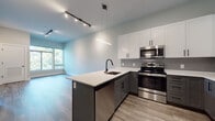 This contemporary single-floor unit showcases a thoughtfully designed open-concept layout that seamlessly connects the kitchen and living area. The kitchen is a standout feature, offering a two-tone cabinet design with crisp white upper cabinets and charcoal gray lower cabinets, all accented by sleek matte black hardware. A generous peninsula island with a white quartz countertop and an undermount sink with a matte black gooseneck faucet anchors the space, providing both functionality and style. Stainless steel appliances — including an electric range, built-in microwave, dishwasher, and refrigerator — complete the well-appointed kitchen. A striking linear pendant light and a track lighting system overhead add a modern industrial touch throughout the open living area. Durable wood-look plank flooring runs throughout the entire unit, adding warmth and continuity. The bedroom is bathed in natural light through large windows that frame lush, green tree views, creating a peaceful retreat. The bathroom features a tub-shower combination enclosed by a bold matte black-framed sliding glass door, complemented by matching black fixtures and clean white finishes. The overall aesthetic is modern, clean, and cohesive, with a neutral color palette that lends itself well to personalization.