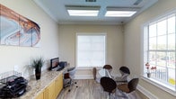 This single-floor space is thoughtfully designed to support both individual work and collaborative gatherings. The main area serves a dual purpose as a business center and casual meeting room, combining practicality with a clean, modern aesthetic. A built-in granite-topped workstation with warm wood cabinetry lines one wall, providing ample desk space complete with a computer and printer — ideal for administrative tasks or remote work needs. Overhead, a set of flush-mounted skylight panels flood the room with bright, even artificial light, creating a welcoming and energized atmosphere. The neutral beige walls, accented by crisp white crown molding and baseboards, lend the space a polished and professional character. Light-toned hardwood-style flooring runs throughout, adding warmth and visual continuity. At the center of the room, a round glass-top table surrounded by modern curved chairs in dark tones offers an inviting spot for small group meetings or casual conversations. A window dressed with white horizontal blinds allows for natural light and privacy control, while a decorative plant near the workstation adds a subtle touch of greenery. Overall, the space strikes a balance between functionality and comfort.
