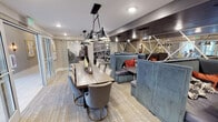 This thoughtfully designed amenity space functions as a multifunctional co-working and social lounge spread across a single floor. The layout is divided into distinct zones that seamlessly complement one another, creating an environment suited for both focused work and casual gathering.

At the heart of the space sits a large, rustic wood communal dining table surrounded by quilted upholstered chairs, suspended beneath a striking black multi-pendant industrial chandelier with gold-lined shades. This centerpiece area is ideal for group meetings or collaborative work sessions.

Flanking the main table is a series of plush blue velvet booth seating units with dark laminate tabletops, designed to provide semi-private nooks for individuals or small groups. The booths are accented with orange and red throw pillows, adding vibrant contrast to the cool-toned palette. A full mirrored wall with geometric diagonal framing lines one side, visually expanding the space and reflecting light throughout.

Dedicated workstations equipped with desktop computers and articulating task lamps provide additional focused work areas. Glass sliding doors and large glass panel walls allow natural light to filter in from an adjacent corridor or lobby, maintaining an open and airy feel. Luxury vinyl plank flooring with a patterned area rug grounds the space, while large-scale black-and-white cityscape photography murals serve as sophisticated decorative backdrops.