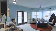 This single-floor living space is defined by its soothing blue walls, which create a calm and welcoming atmosphere throughout. The living area is generously proportioned, anchored by plush carpeting and a warm terracotta-toned circular area rug that adds a grounding contrast to the cool color palette. A ceiling fan with an integrated light fixture provides both comfort and functional illumination overhead, while an additional pendant light enhances the room's brightness.

Natural light is a standout feature, flowing in through a set of dark-framed French doors that offer outdoor access and views, as well as a large window dressed with white sheer curtains and horizontal blinds. Together, these openings lend the space an airy, open quality. The seating area is comfortably arranged with a neutral-toned sofa accented by decorative pillows and a dark wood coffee table, creating an inviting focal point. A side table with a table lamp adds a warm, layered lighting element. Adjacent to the main seating area, a dining surface is partially visible, suggesting a fluid, open-concept layout well-suited for everyday living and entertaining.