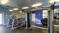 This fitness center is a thoughtfully designed workout facility that provides everything needed for a comprehensive exercise routine. The space features a dark rubberized floor, ideal for high-impact activities, and is brightened by fluorescent lighting and ceiling fans that help maintain a comfortable atmosphere during workouts.

The room is appointed with a solid selection of cardio equipment, including an elliptical trainer and additional aerobic machines, all reflected in the full-length wall mirrors that enhance the sense of space and allow users to monitor their form. A Life Fitness cable machine serves as a centerpiece for strength training, complemented by a well-stocked dumbbell rack along the far wall, offering a range of free weights for users of all fitness levels.

A bold blue accent wall adds a dynamic, energetic visual element to the space, while a posted rules board near the entry helps facilitate organized use of the facility. A secondary area visible beyond the main floor appears to offer additional equipment or a stretching zone, adding versatility to the overall layout. The single-floor layout ensures easy accessibility, making this a practical and motivating environment for daily fitness.