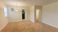 This single-floor residence at 204 D offers a straightforward and functional layout with a calm, neutral aesthetic throughout. The main living area is finished with wall-to-wall carpeting in a warm beige tone, providing a soft and cohesive foundation across the space. Walls are painted in a light, neutral shade that contributes to an airy and open feel.

A notable architectural detail is the arched doorway that connects the main living area to an adjacent room, adding a subtle classic charm to the otherwise simple design. A pendant light fixture with a clean, triangular silhouette hangs overhead, serving as a modest focal point in the dining or central area.

A doorway leads to what appears to be a kitchen, where dark cabinetry and a black refrigerator are visible, offering a contrast to the lighter tones found in the rest of the space. White baseboards run consistently throughout, providing a clean finish along the floor line.

The overall ambiance is understated and move-in ready, with a neutral palette that allows for flexible furnishing and personalization.